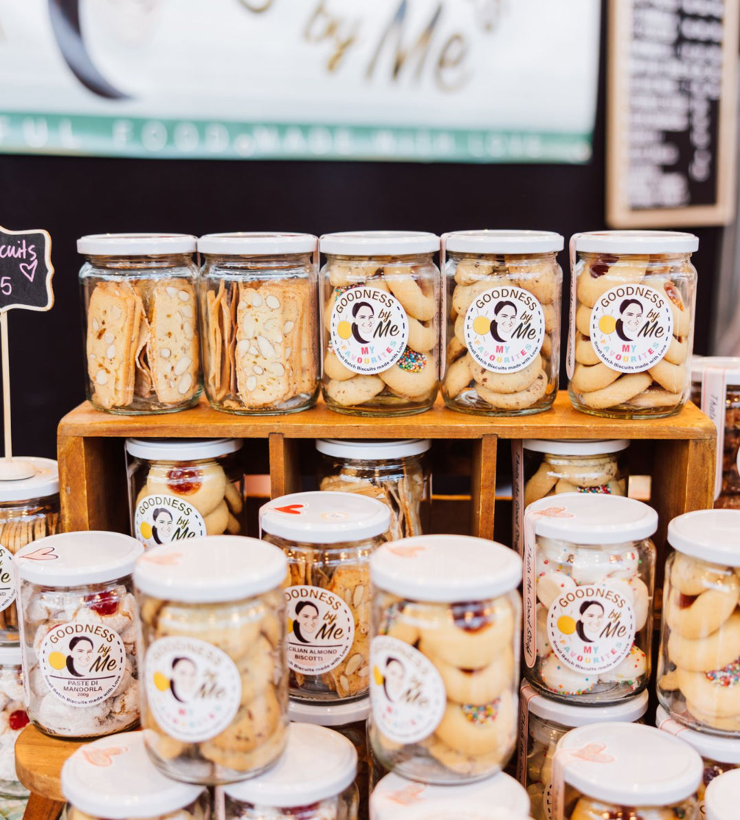 Jars of cookies with 'Goodness Me' labels on a wooden shelf.