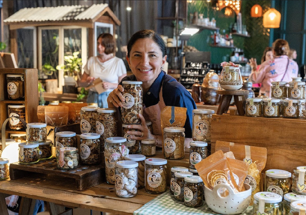 Linda from Goodness by Me holding a jar of granola in a market setting with various jars and products displayed.